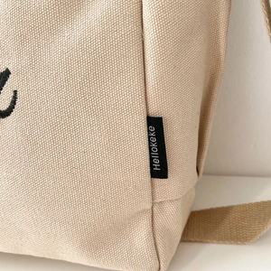 Detail showing the textured canvas material and black logo tag on a beige tote bag