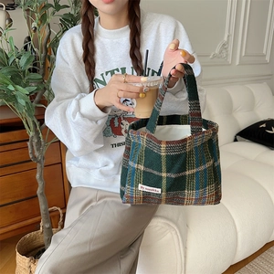Detail of green plaid wool tote bag with white tag held by model in home setting