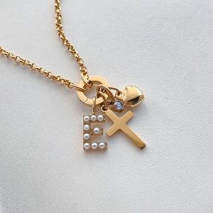 Dettaglio macro di una collana in acciaio al titanio oro con croce e iniziale di perle.