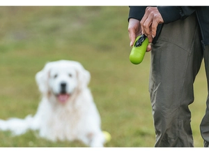 Green pill-shaped pet waste bag dispenser being used on a lawn with a blurred dog