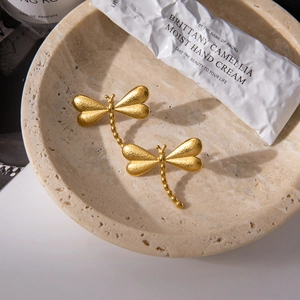 Gros plan de boucles d'oreilles libellule en or 18K mat avec texture brossée sur marbre