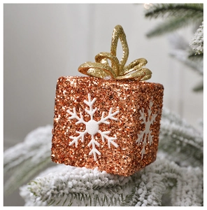 Close-up of golden glitter gift box ornament with white snowflake on snowy pine branch