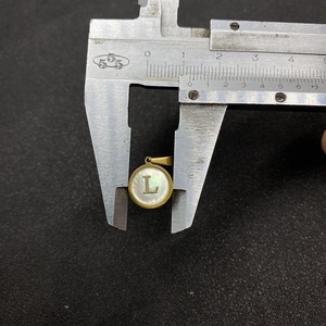Close-up of a golden shell pendant with letter L being measured by a caliper