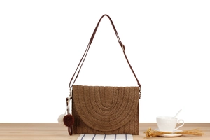 Detail of a brown woven paper straw crossbody bag featuring decorative plush pom-pom charms