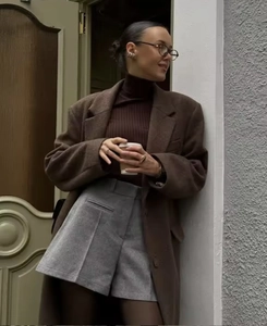 Street style shot of a woman in a brown wool coat and grey shorts holding a coffee cup
