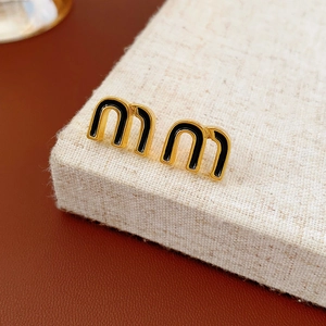 Detail of gold letter M earrings with black enamel inlay on beige textured surface