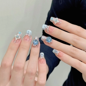 Nahaufnahme von langen Kunstnägeln mit blauem Glitzerverlauf und 3D-Blumen-Edelsteinen