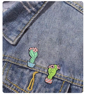 Close-up of two cartoon cactus enamel pins on a denim jacket pocket edge with pink flowers