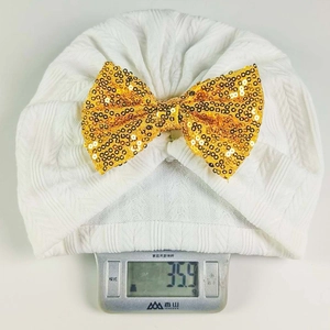 Detailed weight measurement showing white baby turban hat with golden sequin bow on scale