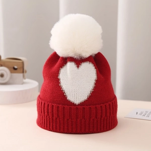 Close-up of red knit infant hat with white faux fur pompom and silver heart embroidery