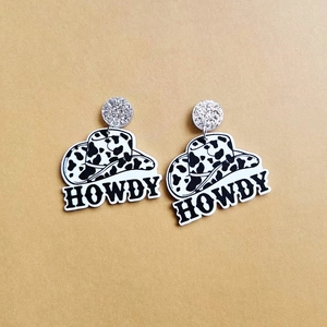 Close-up of cow print cowboy hat earrings with black acrylic HOWDY letter charms