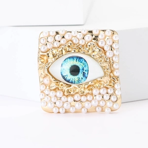 Detail of square gold brooch with white pearls and blue evil eye centered on white cube
