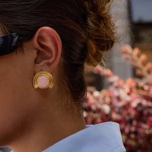 Detail of a gold sunburst stud earring with a pink stone worn by a model in nature