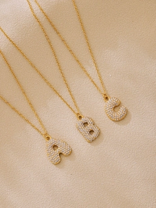 Detail of gold zircon initial necklaces featuring A, B, and C letters on beige surface
