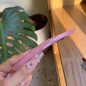 Minimalist pink acetate hair clip with gold logo held against a natural home backdrop