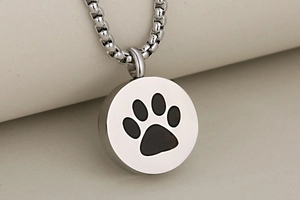 Close-up of a silver stainless steel round cremation pendant with a black paw print design
