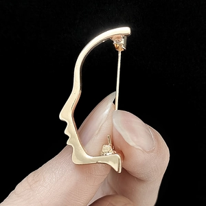 Detailed view of a gold-toned artistic face profile brooch held by fingers on black background