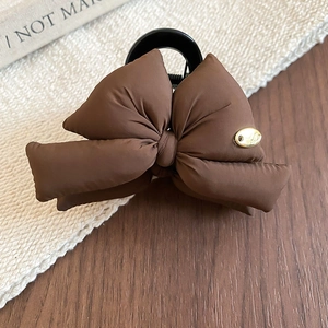 Detail of dark brown padded bow hair claw clip with a small gold logo on a textured backdrop