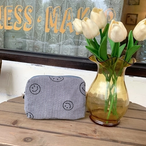 Grey corduroy makeup bag with smiley face embroidery on a rustic wooden table detail