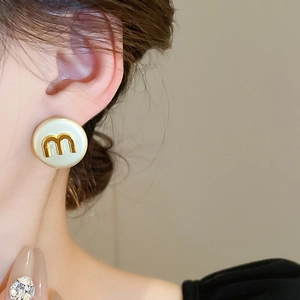 Macro detail of round cream earring with a golden metal m-shaped letter logo on model