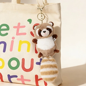 Cute raccoon plush keychain with striped long tail hanging on a beige canvas bag