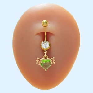 Piercing per ombelico dorato con gemma a cuore verde e ali su sfondo blu chiaro