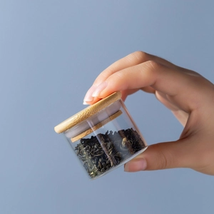 Hand holding portable mini glass tea canister with airtight bamboo lid on grey background