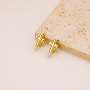 Detail view of golden cross stud earrings on a textured stone over pink background