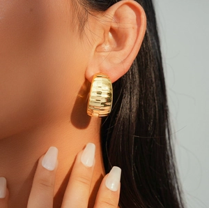 Detail of gold C-shaped earring with horizontal stripes on model with white nails