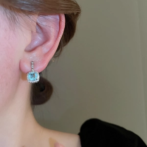 Close-up of a square light blue gemstone earring with a diamond halo on a model ear