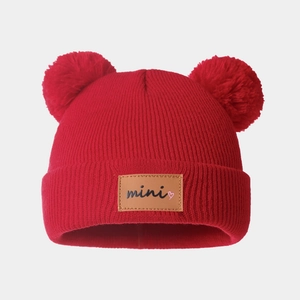 Detail of red knit baby hat featuring double faux fur pom-poms and mini leather patch