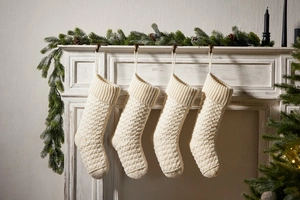 Detail of four cream knitted Christmas stockings hanging on fireplace with pine decor