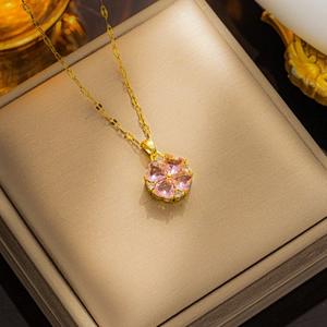 Detailed close-up of a gold clover necklace with pink heart-shaped crystal on a beige box