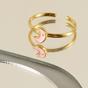 Adjustable gold ring with pink enamel moon and star design on a reflective surface