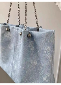 Detail of light blue denim tote bag featuring silver chain straps and metallic accents