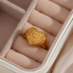 Macro detail of golden heart signet ring engraved with SELF LOVE CLUB in pink velvet box