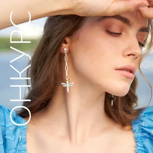 Close-up of a minimalist silver dragonfly pendant earring worn by a model near the water