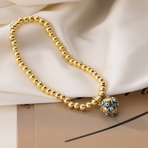 Gold beaded bracelet featuring evil eye enamel pendant on white silk and earth-tone cards