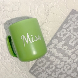 Detail of glitter silver Miss sticker on a matte green mug with DIY alphabet sheet