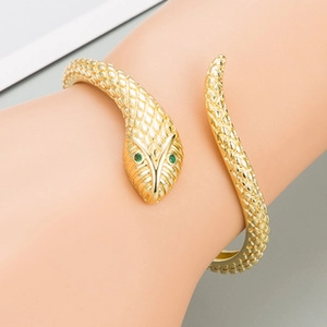 Detail of gold snake-shaped bangle with scale texture and green gemstone eyes on wrist