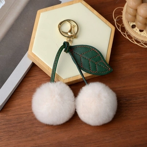 Macro detail of plush cherry keychain featuring cream faux fur balls and green leather leaf