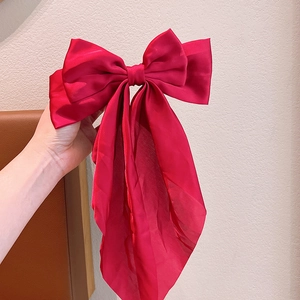 Large red satin bow hair clip with long ribbons shown held in hand against a neutral background