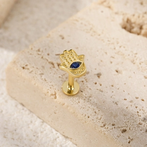 Macro view of a gold Hamsa hand piercing stud with a blue eye stone on a stone background