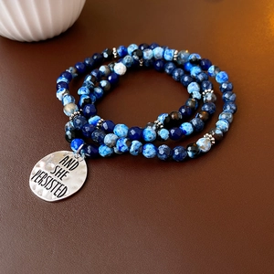 Macro shot of blue beaded multi-layer bracelet with AND SHE PERSISTED engraved pendant