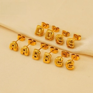 Detail view of five polished gold letter earrings A through E on a beige fabric background