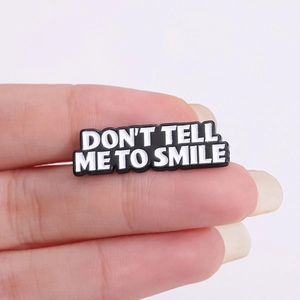Metal badge pin with 'DON'T TELL ME TO SMILE' text held on fingers over white background