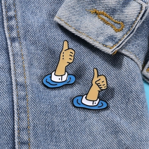 Two cartoon thumbs up enamel pins displayed on a denim jacket collar for fashion
