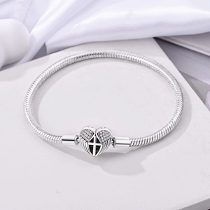 Detail of silver snake chain bracelet featuring heart clasp with cross and wings