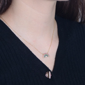 Macro detail of a gold necklace featuring a semi-circle rainbow pendant with multi-color stones