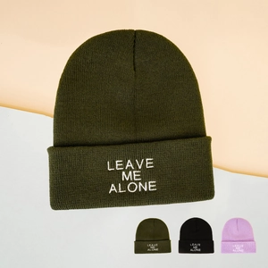 Close-up of white LEAVE ME ALONE embroidery on a dark green knitted beanie hat